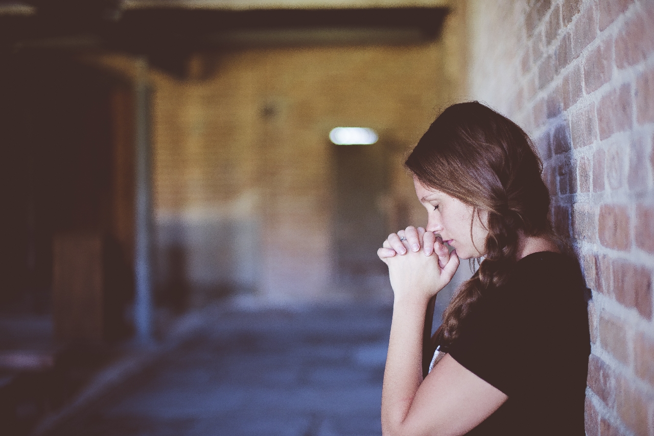 a girl sitting praying with her hands clasp and eyes closed sitting in a alley way