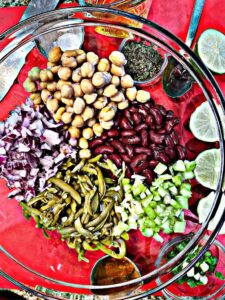 in a glass clear bowl we have kidney beans, garbanzo beans or chick pea, green beans, celery and onion, on a read cutting boardwe have limes, parsley, olive oil salt