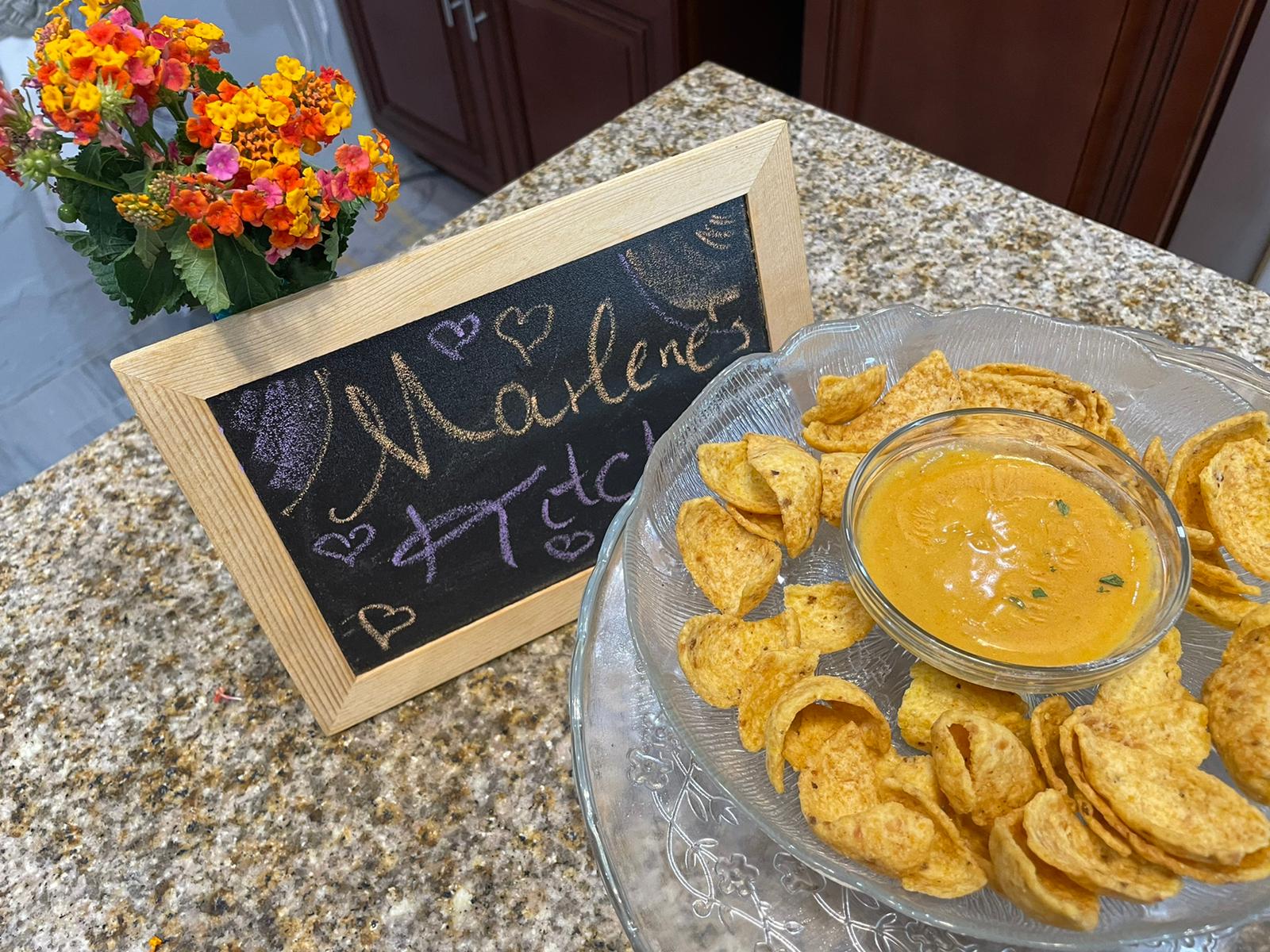 glass bowl of cheese with corn chips around, a bunch of wild flowers and a chalkboard that says Marlene's Kitchen