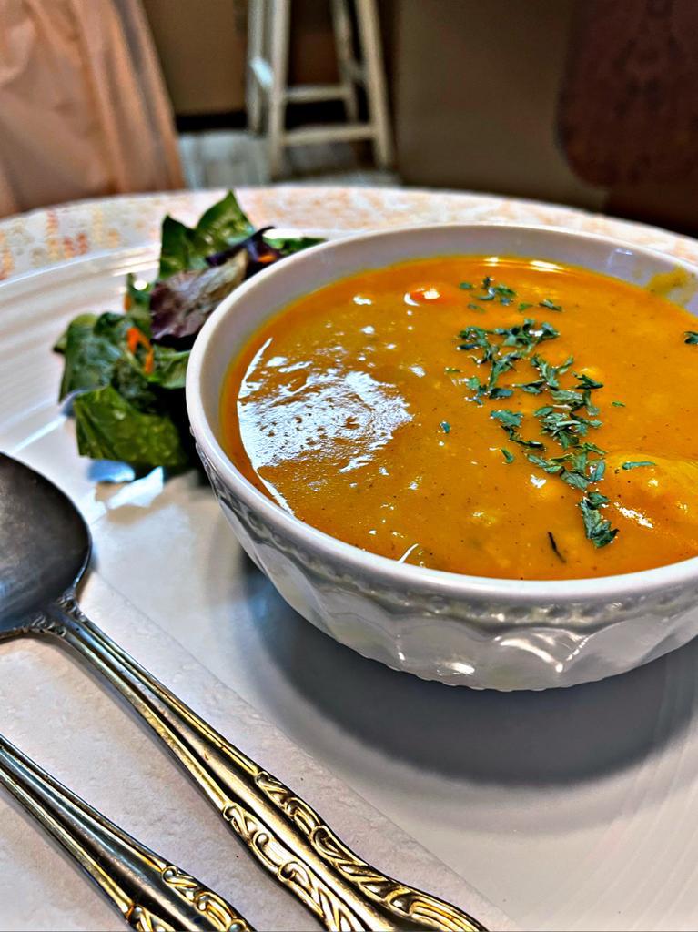 a bowl of pumpkin soup on a small table with spoon and fork on a plate with a green salad