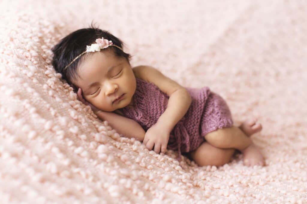 a baby girl in a purple dress asleep on a pink bedding