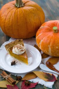 2 large pumpkins with a slice of pumpkin pie on a white small plate and fall leaves