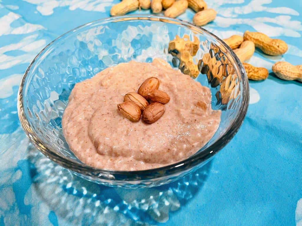 a bowl of peanut porridge with peanuts on top and shelled peanuts on the aqua counter top 